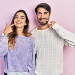 Young Hispanic couple wearing casual clothes smiling cheerfully and pointing with fingers at their perfect teeth.