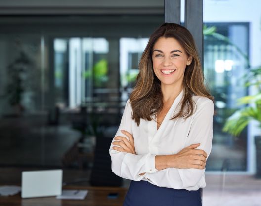 A cheerful confident young businesswoman with a perfect smile standing with folded arms at the desk in her office