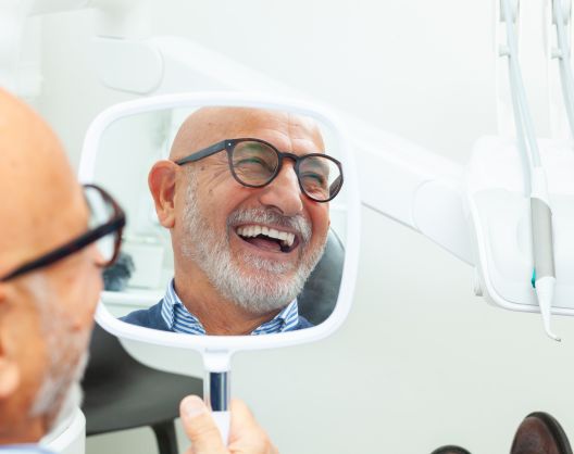A smiling older man with glasses and a beard looks at his reflection in a handheld mirror while sitting in a dental office.