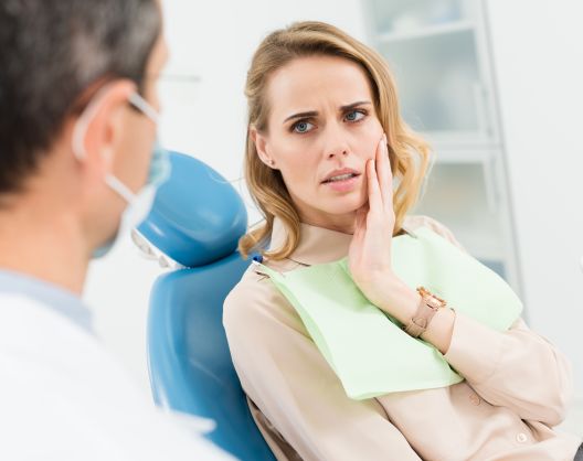 A concerned woman sits in a dental chair, touching her cheek in pain, while a dentist in a white coat and mask speaks to her during a dental consultation.