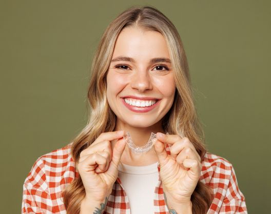 Smiling young woman with long blonde hair, wearing a checkered shirt, holds a clear dental aligner in both hands in front of her face, standing against a plain olive green background.