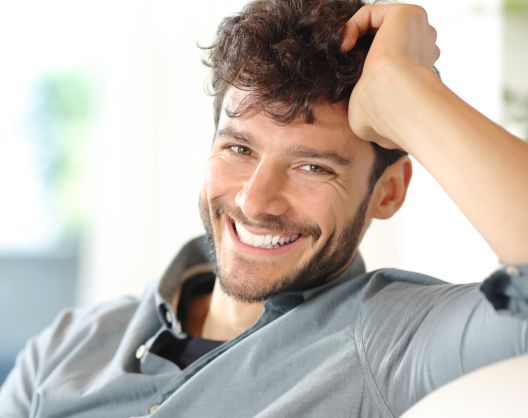A man with short, curly brown hair and a beard sits smiling brightly, leaning his head on his hand. He is wearing a light blue button-down shirt and appears relaxed in a softly lit indoor setting.